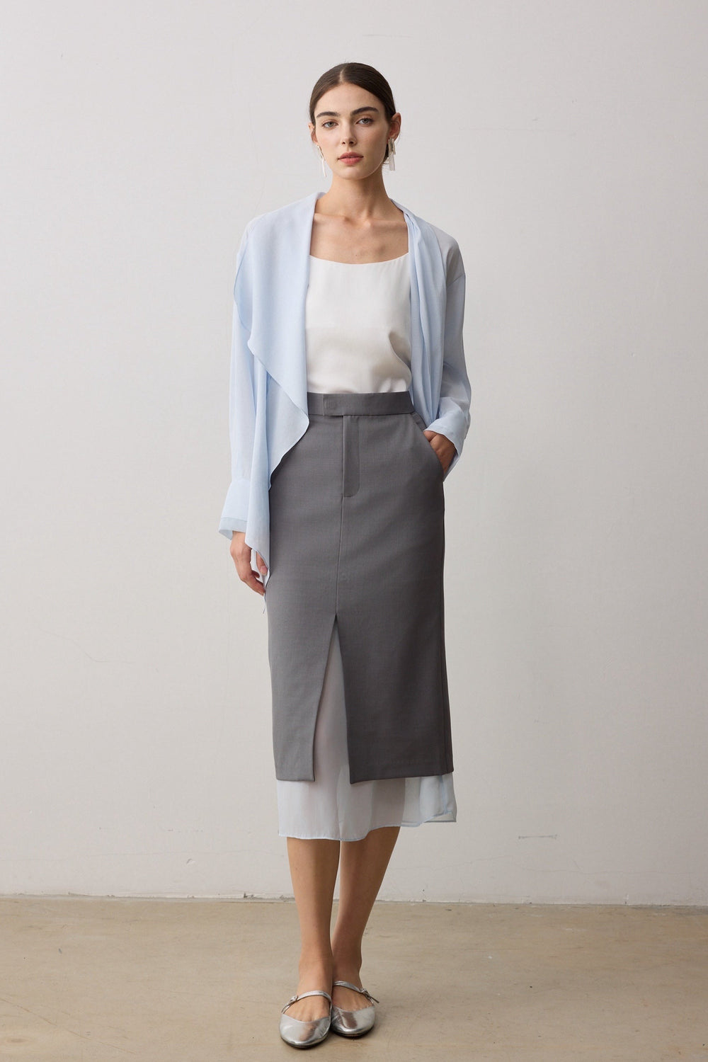 A woman stands in a studio wearing a light blue open-front cardigan over a white top, paired with a high-waisted gray midi skirt and metallic silver flats.
