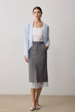 A woman stands in a studio wearing a light blue open-front cardigan over a white top, paired with a high-waisted gray midi skirt and metallic silver flats.